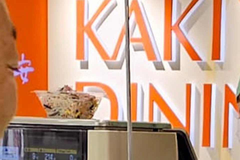 Two people stand at a food counter. One wears a green "TripGuru" shirt; the other wears a hat. Staff in uniform assists. "Kakiyasu Dining" sign in background.