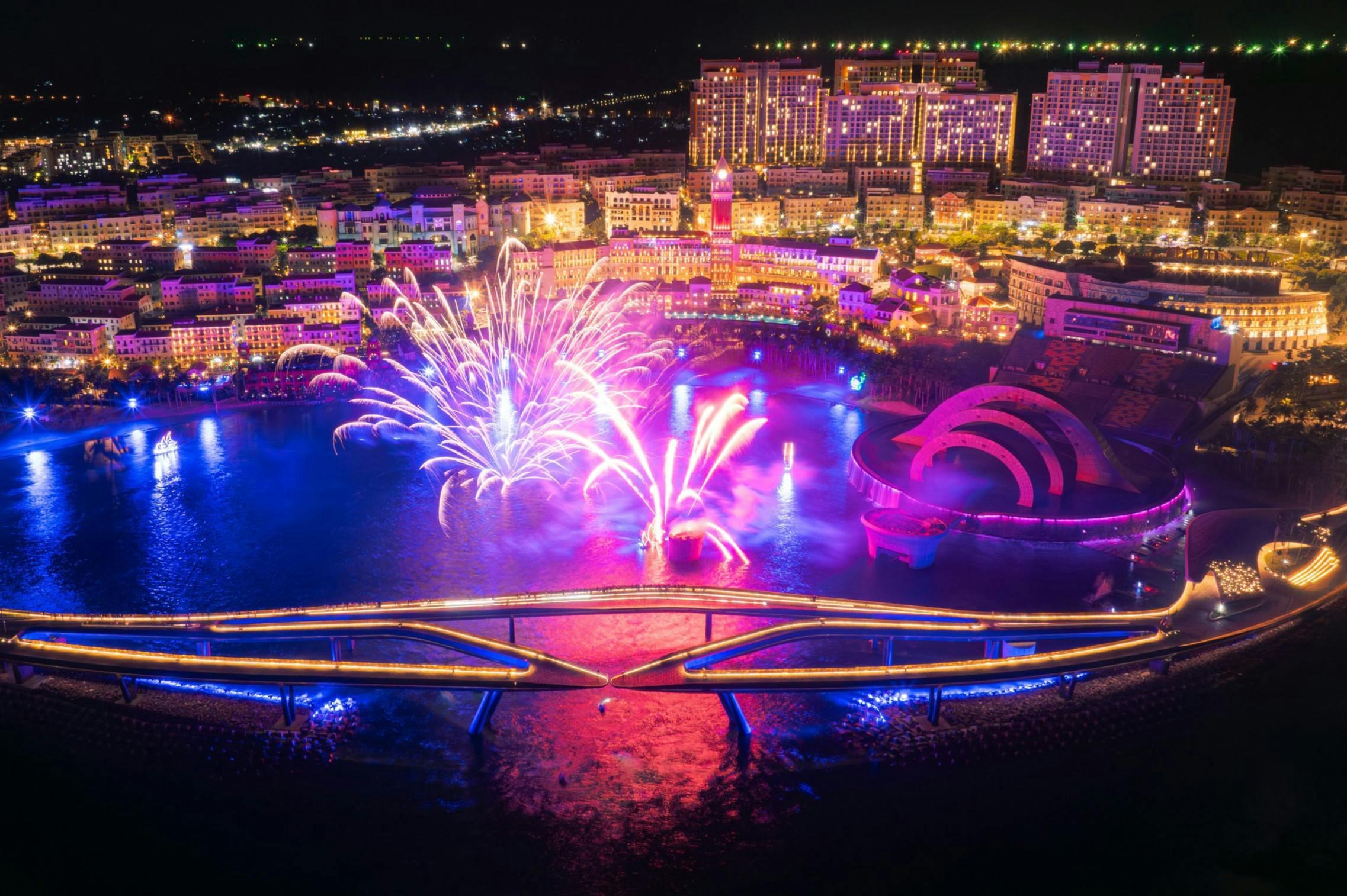 Colorful fireworks over a lit-up waterfront cityscape at night, featuring a modern bridge and illuminated buildings.