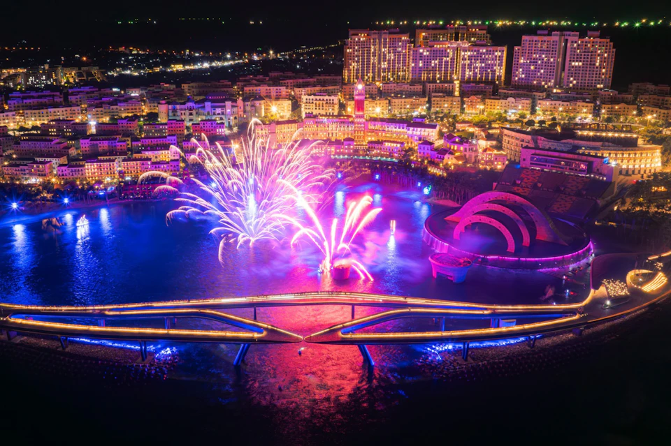 A vibrant nighttime cityscape with colorful fireworks over water, modern architecture, and illuminated bridges.