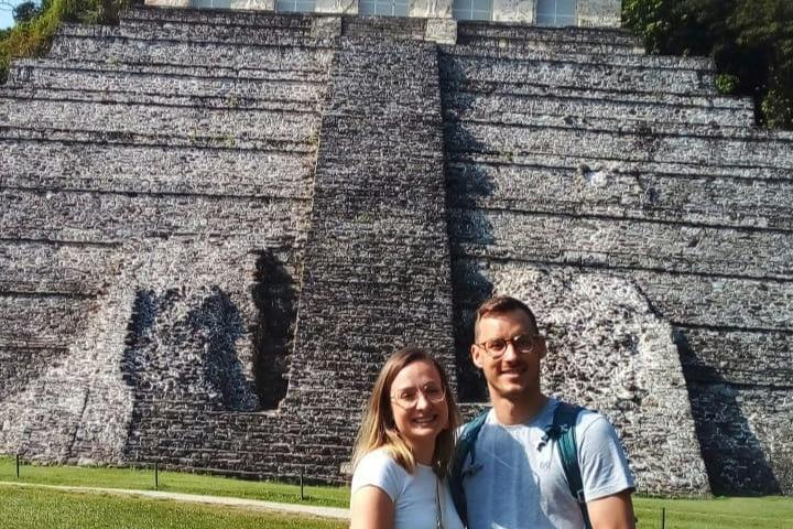 Two people smiling in front of a large stone pyramid structure under a sunny sky.
