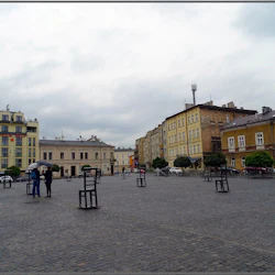 Öppet torg med spridda stora trästolar, omgivna av flervåningshus. Två personer som delar ett paraply i mitten.