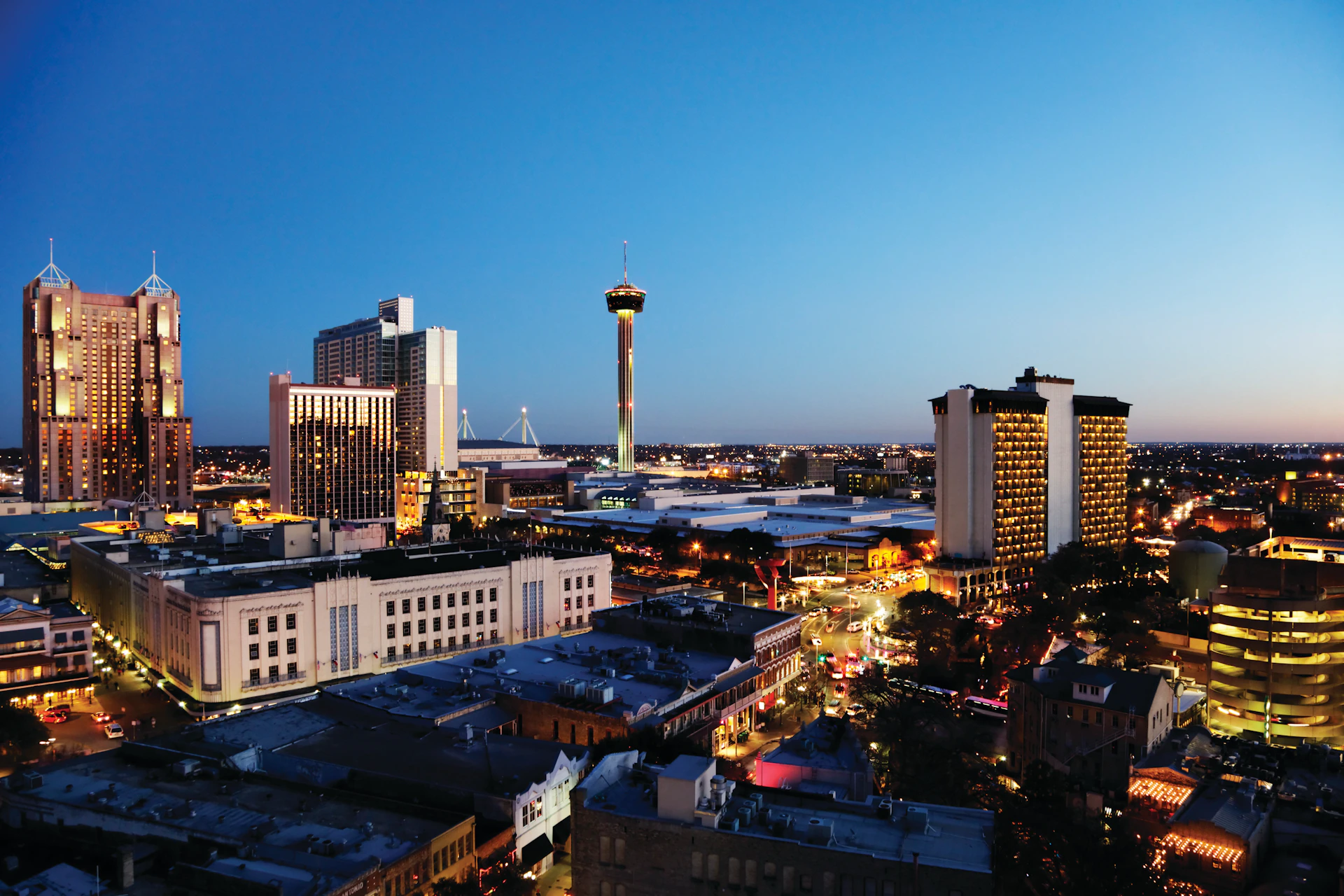 Cityscape at dusk with tall buildings, a prominent tower in the center, and streets lit with car and building lights.
