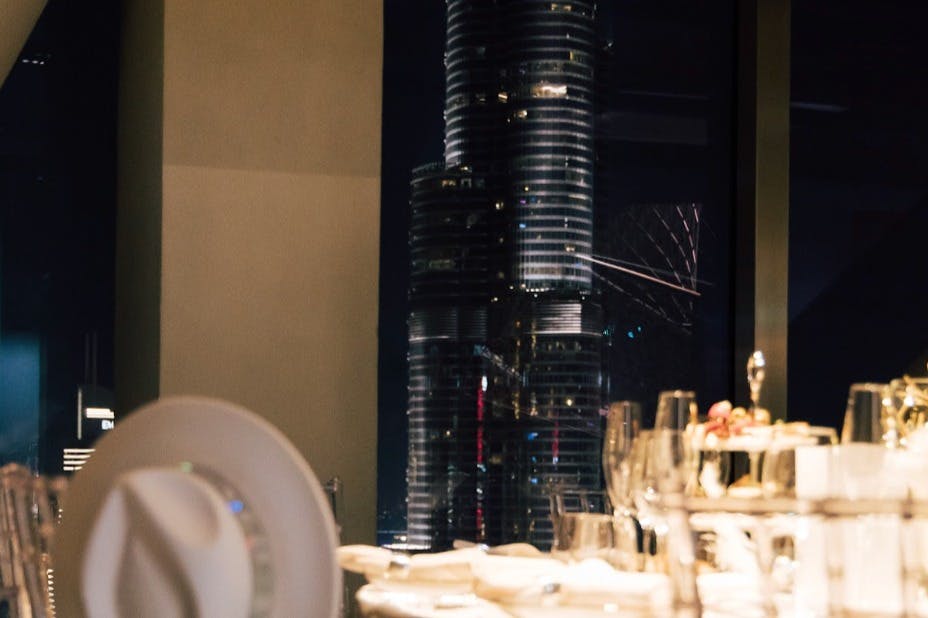 透过窗户可以看到现代摩天大楼的夜间城市景观，前景设有配备玻璃杯的餐桌。