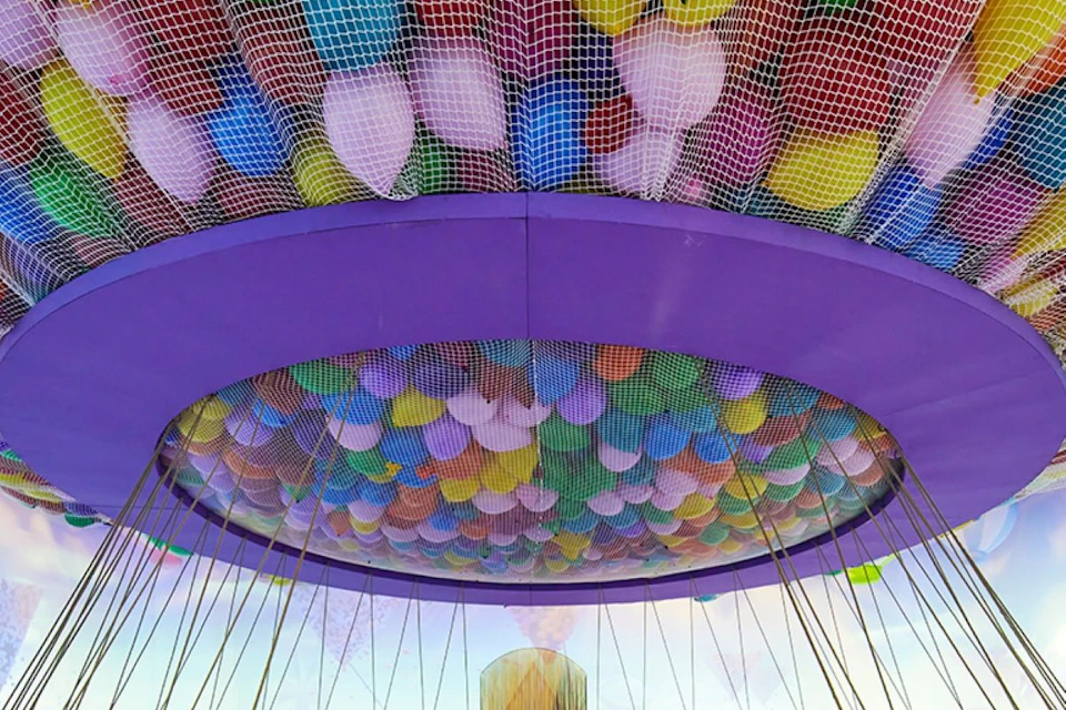 A large cluster of colorful balloons is contained within a net, suspended from a circular structure with a purple rim.