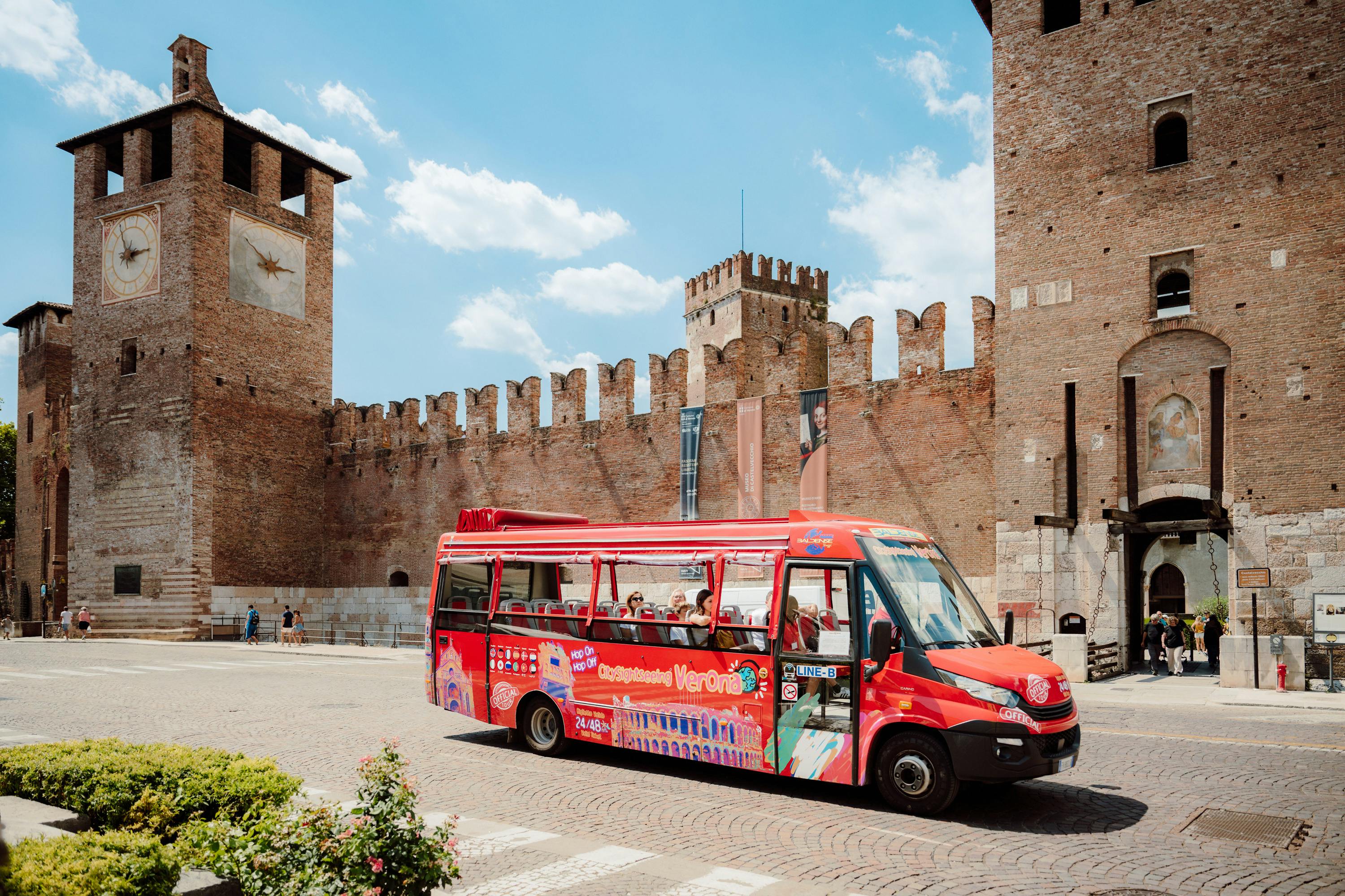 Un autobus turistico rosso davanti alle mura di un castello storico in mattoni sotto un cielo blu con nuvole.