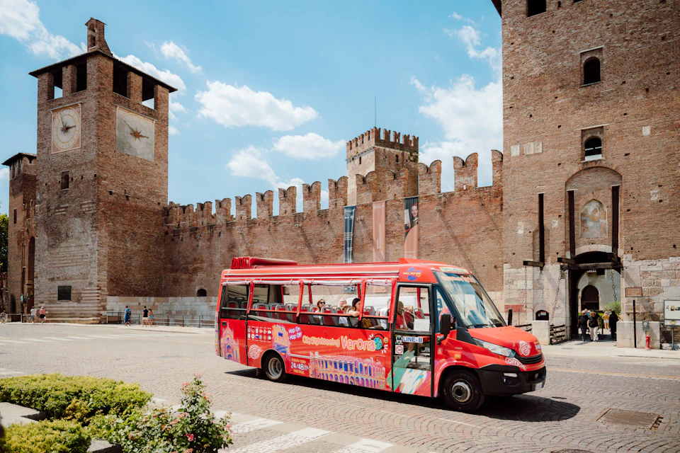 A red tour bus in front of a historic brick castle wall under a blue sky with clouds.