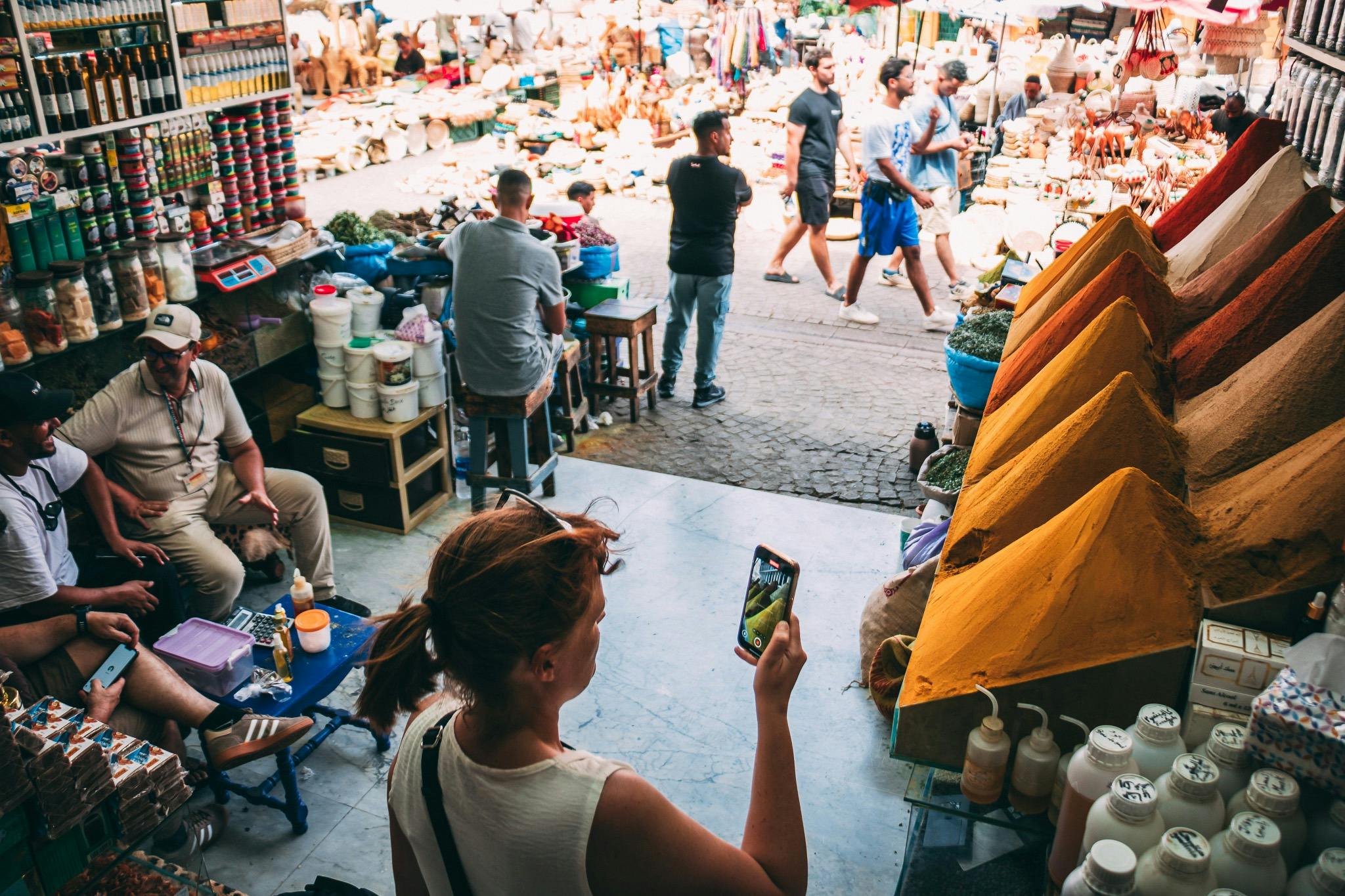 Femme tenant un smartphone dans un marché extérieur animé, avec des gens qui marchent et des marchandises diverses exposées sur des tables.