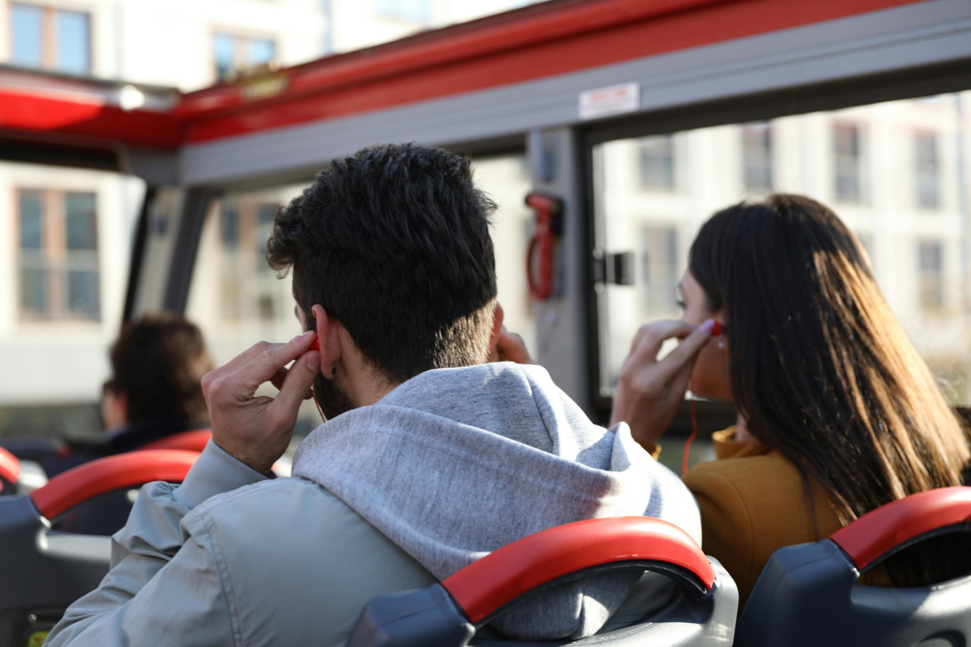 Two people sitting on a bus with red seats, wearing earphones and looking out the window.