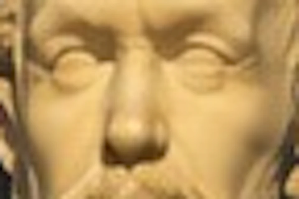 A marble bust of a man with a beard on a black pedestal, placed against a dark blue wall with spotlight illumination.