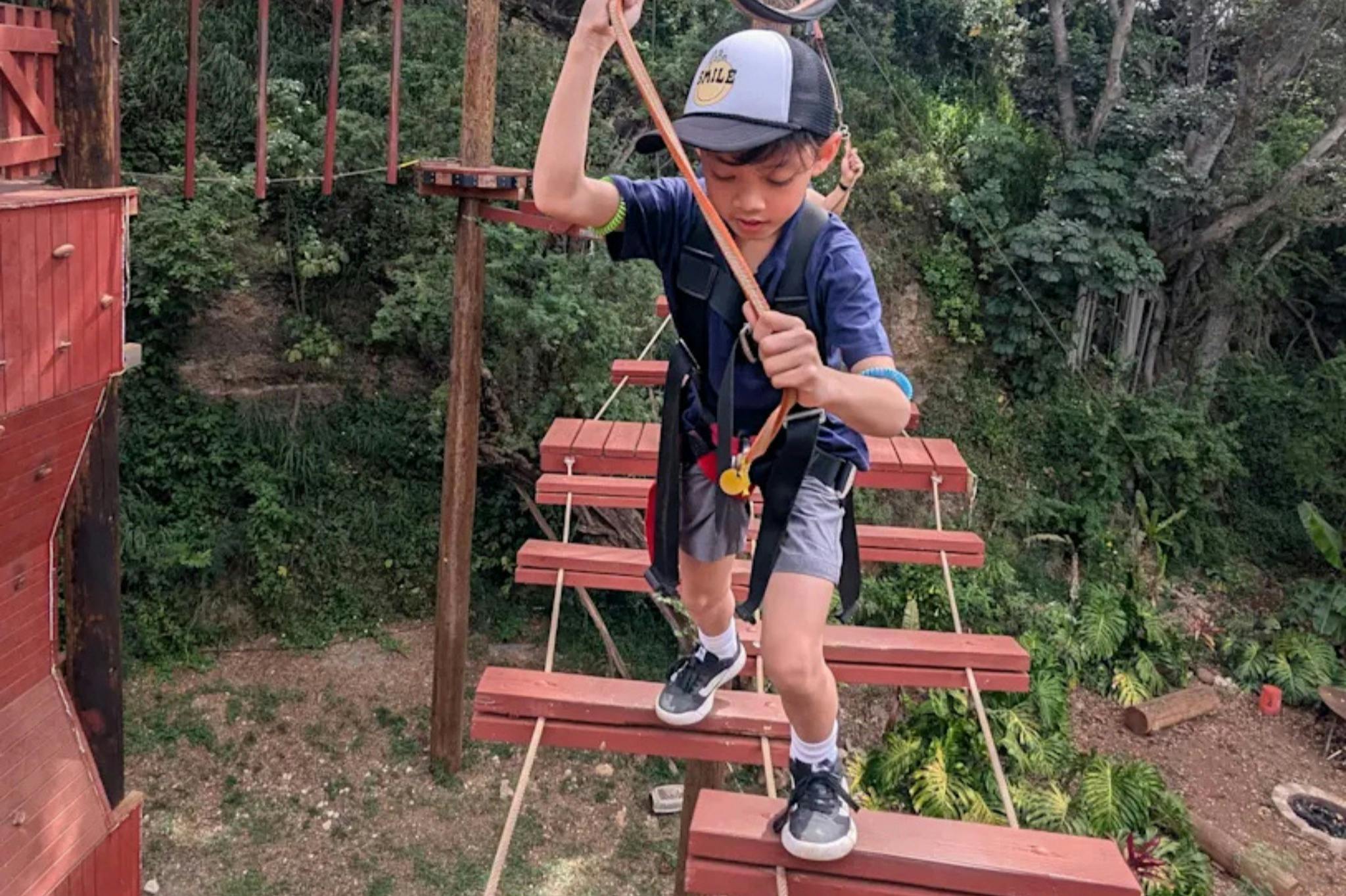 A child wearing a harness crosses a wooden plank bridge in an outdoor ropes course surrounded by greenery.