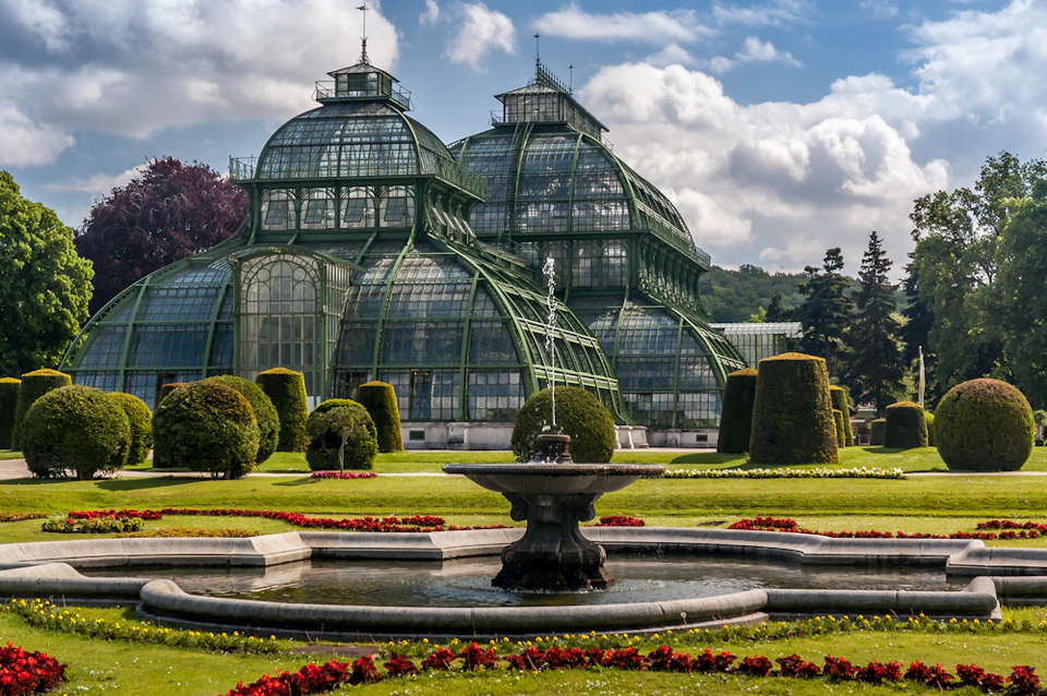 A large glass conservatory with a domed roof. A fountain and manicured gardens are in the foreground under a partly cloudy sky.