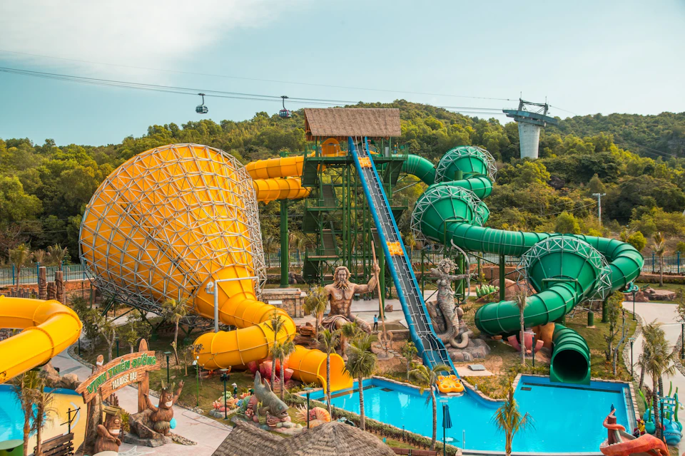 A colorful water park with large yellow and green slides, a pool, and statues, set against a forested hill backdrop.