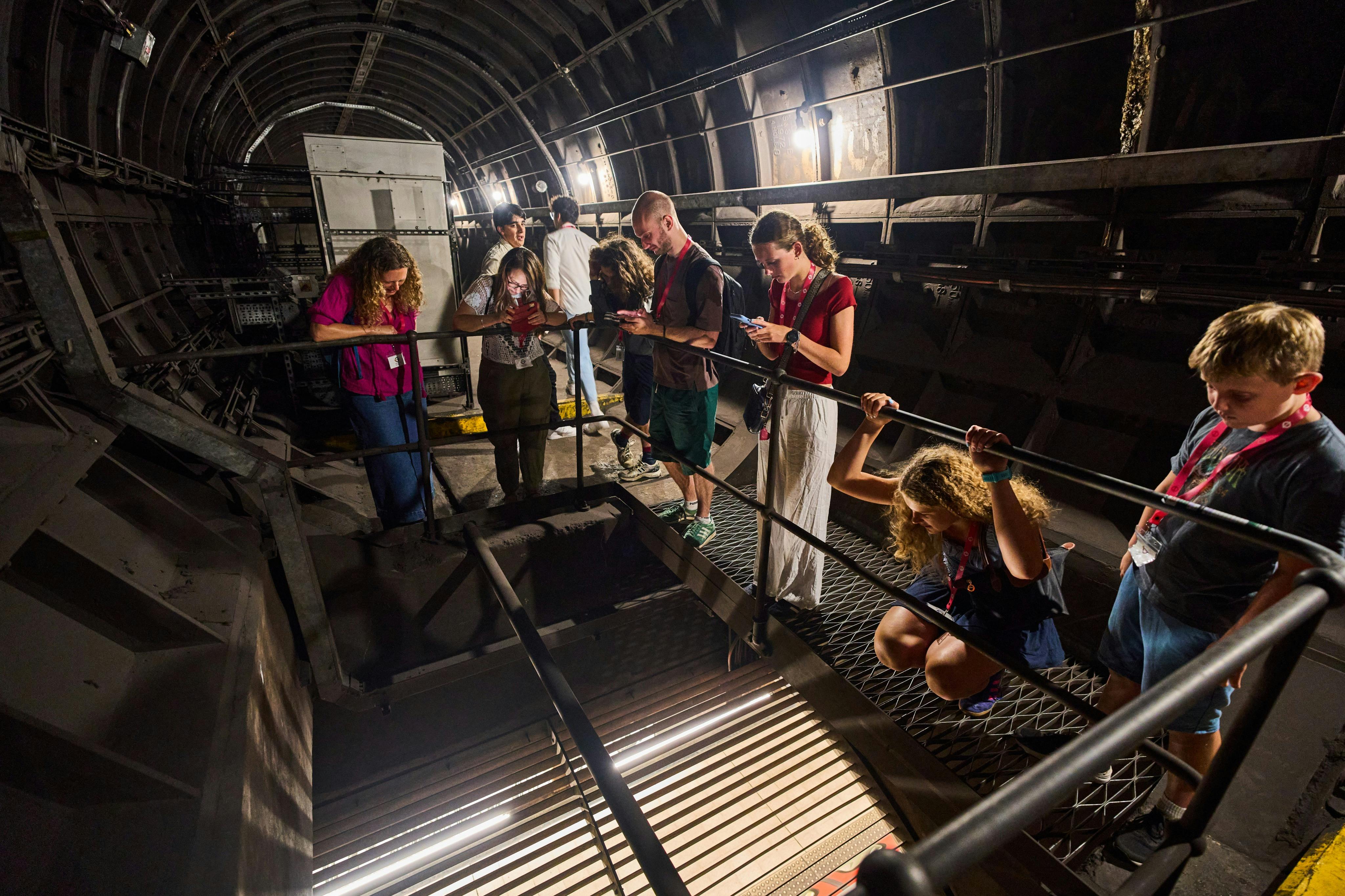 Besucher, die einen unterirdischen Tunnel erkunden, stehen auf Metallstegen, einige schauen nach unten, andere machen Fotos oder beobachten.