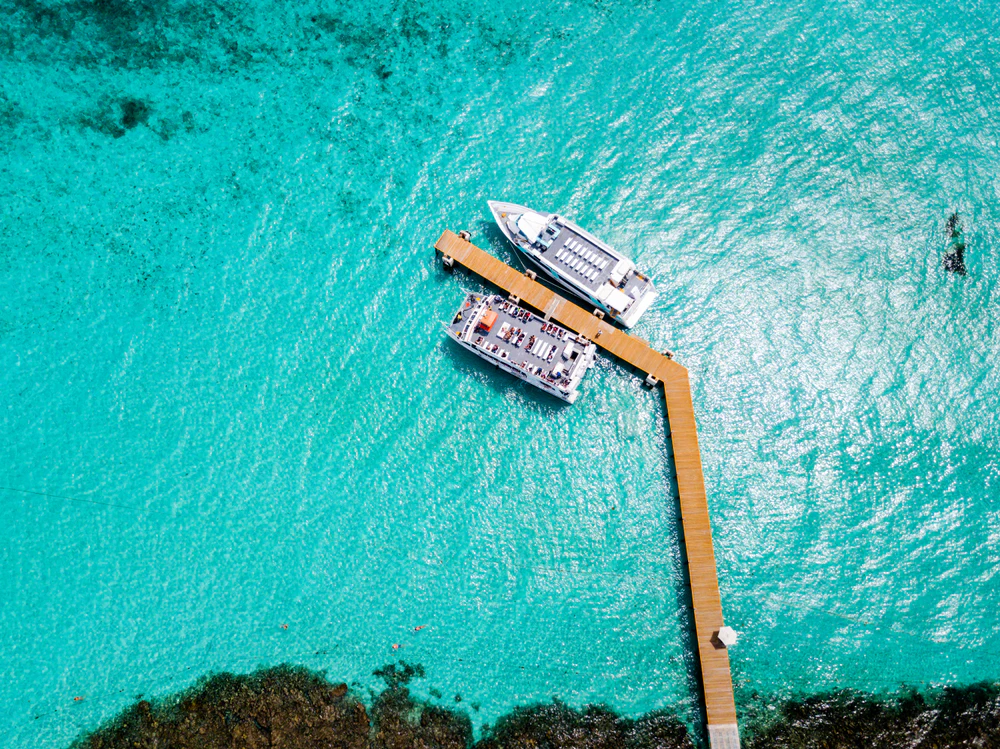 Two boats docked at a wooden pier, surrounded by clear turquoise water with visible patches of underwater reefs.