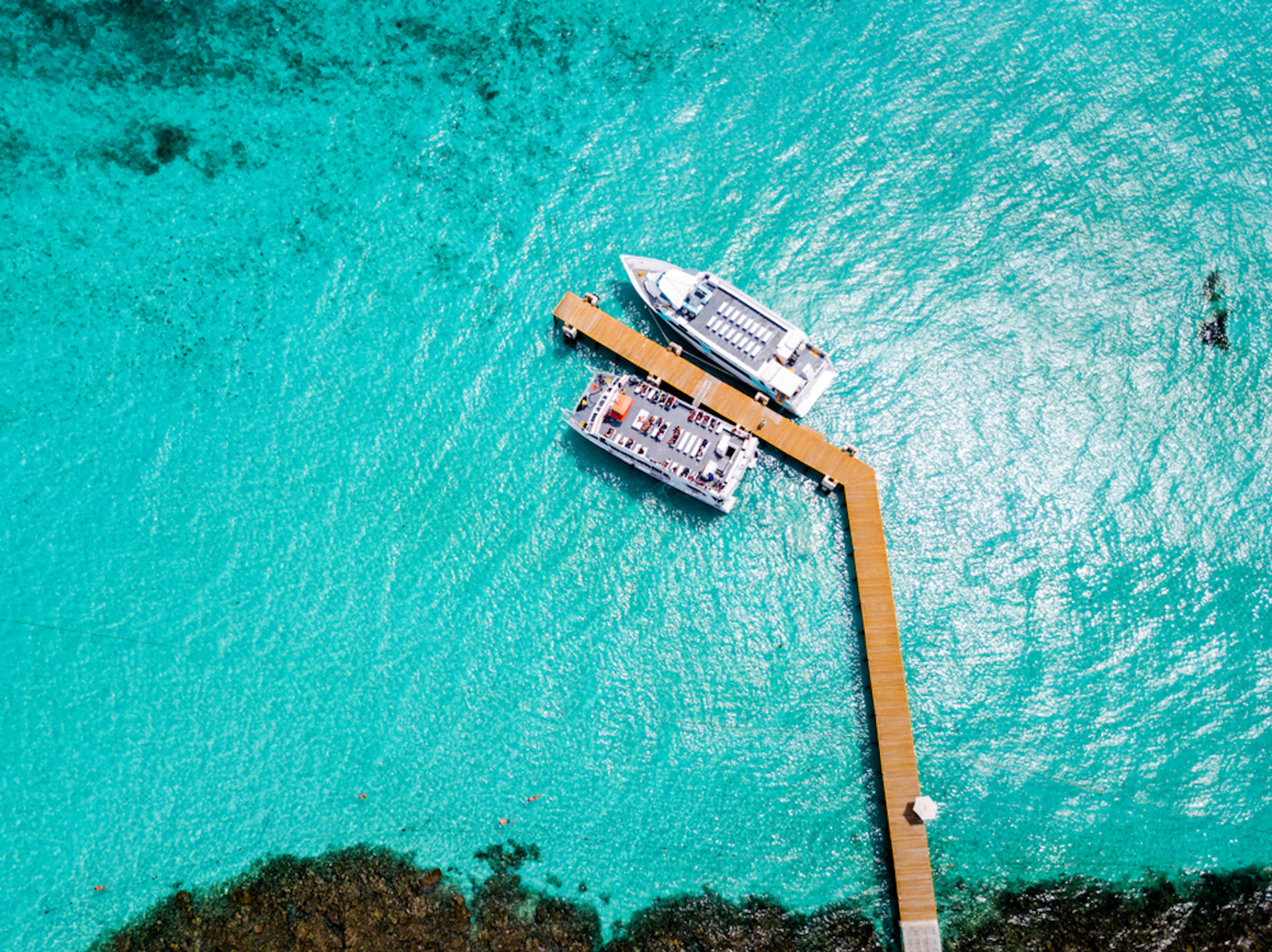 Two boats docked at a wooden pier, surrounded by clear turquoise water with visible patches of underwater reefs.