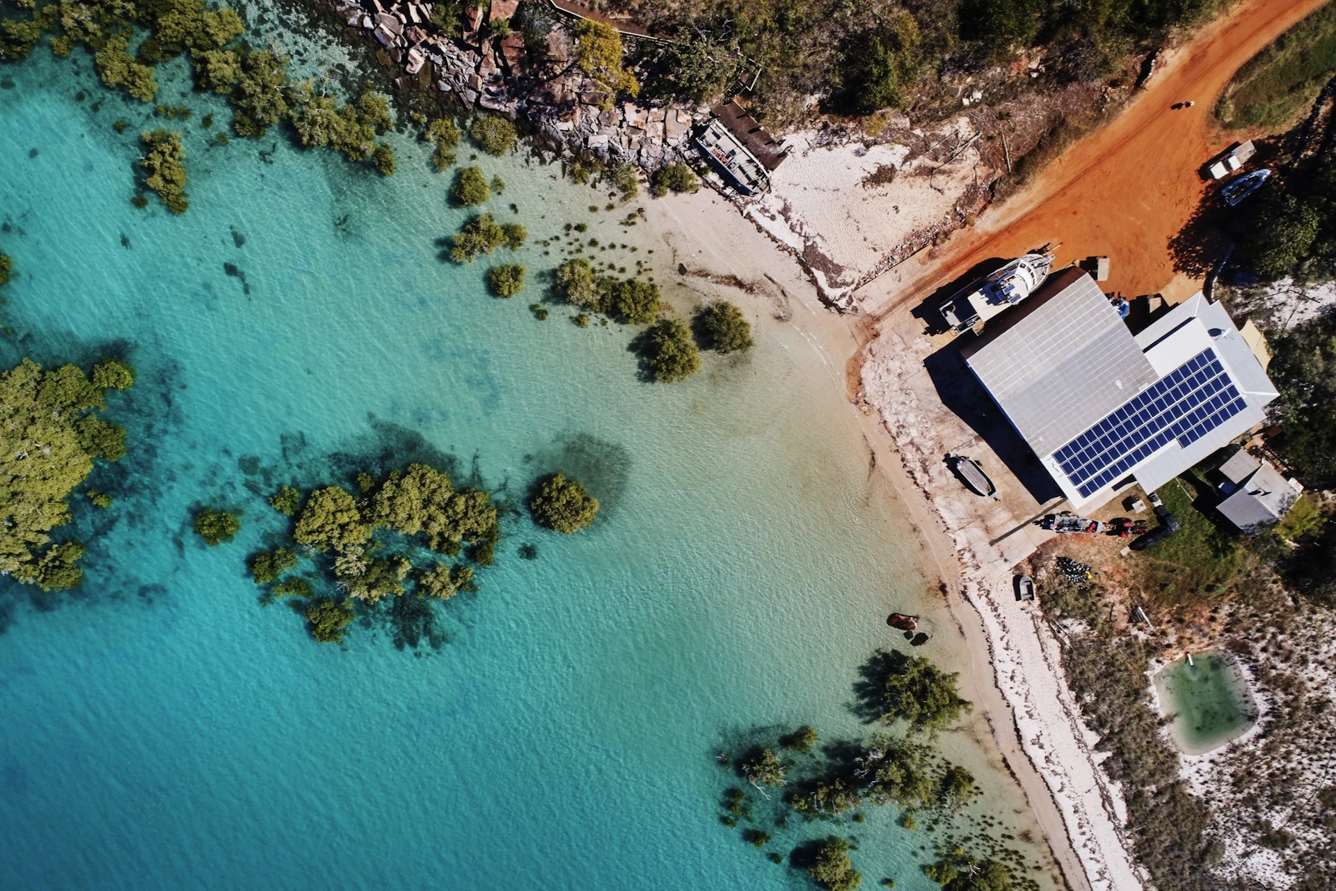 Aerial view of a shoreline with turquoise water, scattered vegetation, a building with solar panels, and a dirt road.