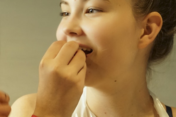 Dwie dziewczyny w plastikowych fartuchach i czerwonych opaskach stoją blisko siebie, uśmiechając się i rozmawiając w pomieszczeniach.