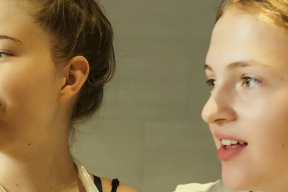Two girls wearing plastic aprons and red wristbands stand close together, smiling and talking indoors.