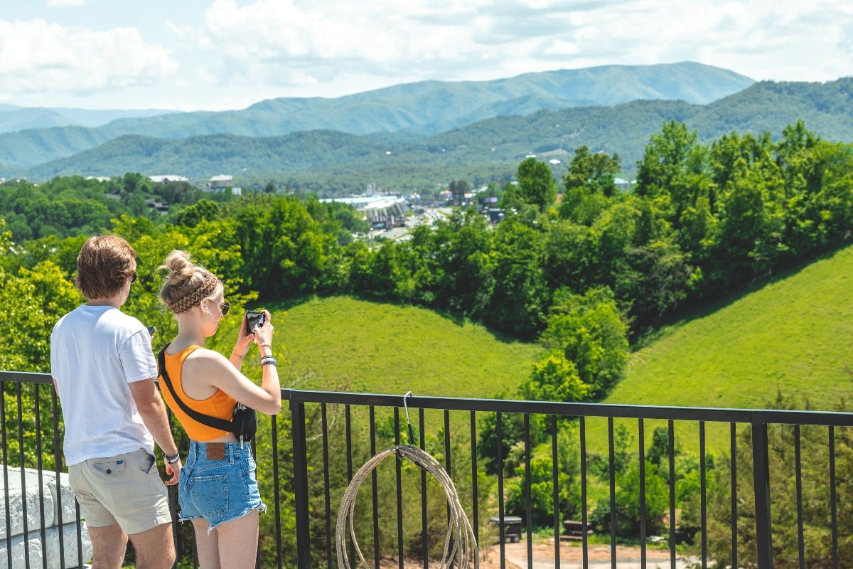 Two people on a balcony overlook a lush landscape with trees and distant mountains. One person holds a camera.