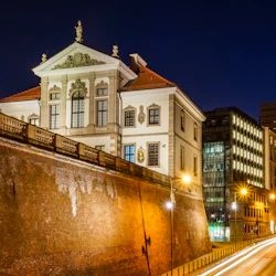 Upplyst byggnad i klassisk stil bredvid en livlig stadsgata, fångad på natten med ljusspår från förbipasserande fordon.