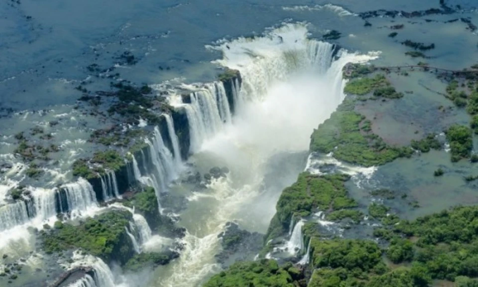 Aerial view of a powerful waterfall with multiple streams cascading into a river, surrounded by lush greenery and rocky terrain.