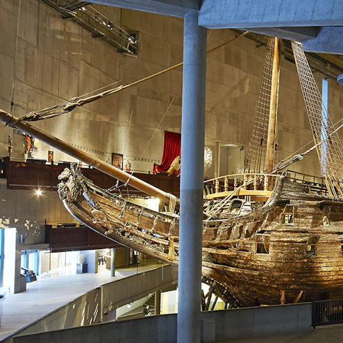 Besökare observerar ett stort, gammalt träskepp som visas i ett rymligt museum med betongväggar och högt i tak.