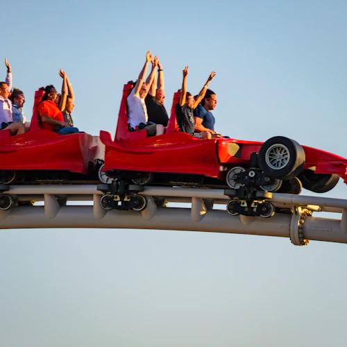 Menschen mit erhobenen Händen fahren in roten Achterbahnwagen, die wie Rennwagen geformt sind, unter einem klaren blauen Himmel.