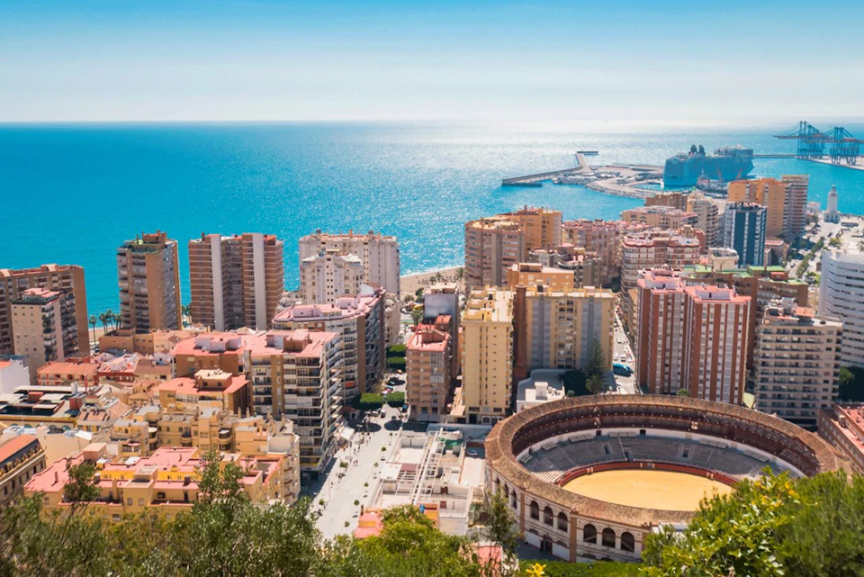 View of a coastal city with high-rise buildings, a circular bullring, and a harbor extending into a calm blue sea under a clear sky.