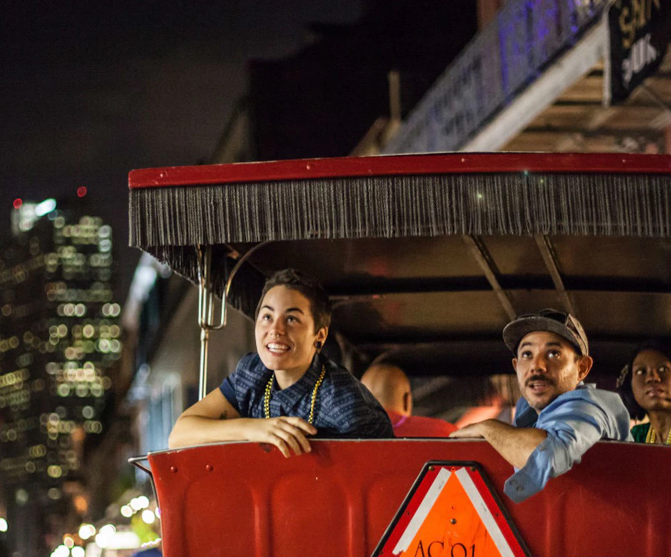 Two people smiling while riding in a red open-air carriage at night, with festive beads around their necks.
