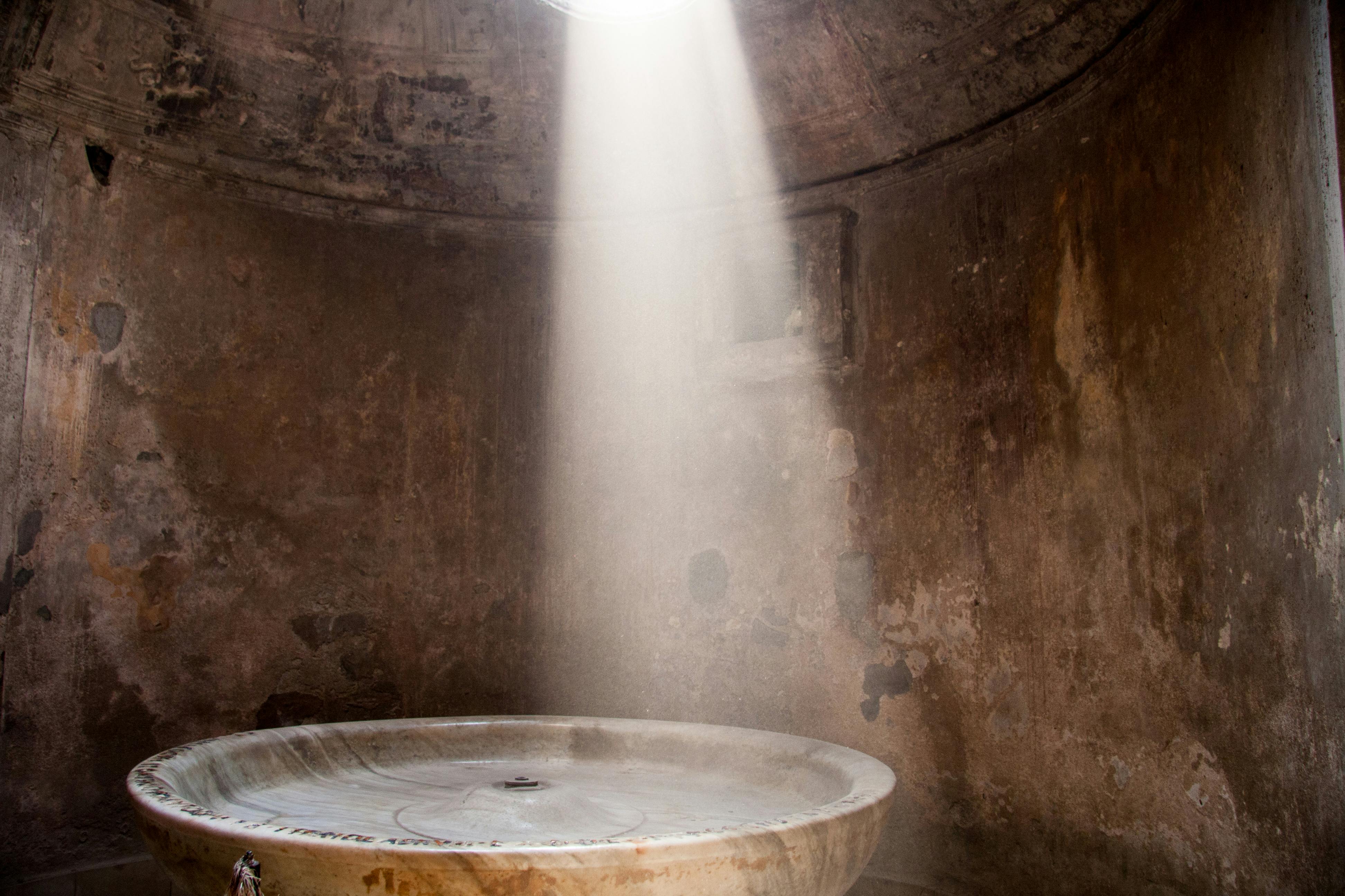 A stone basin stands in an ancient, dimly-lit room with light streaming in from a small hole in the ceiling.
