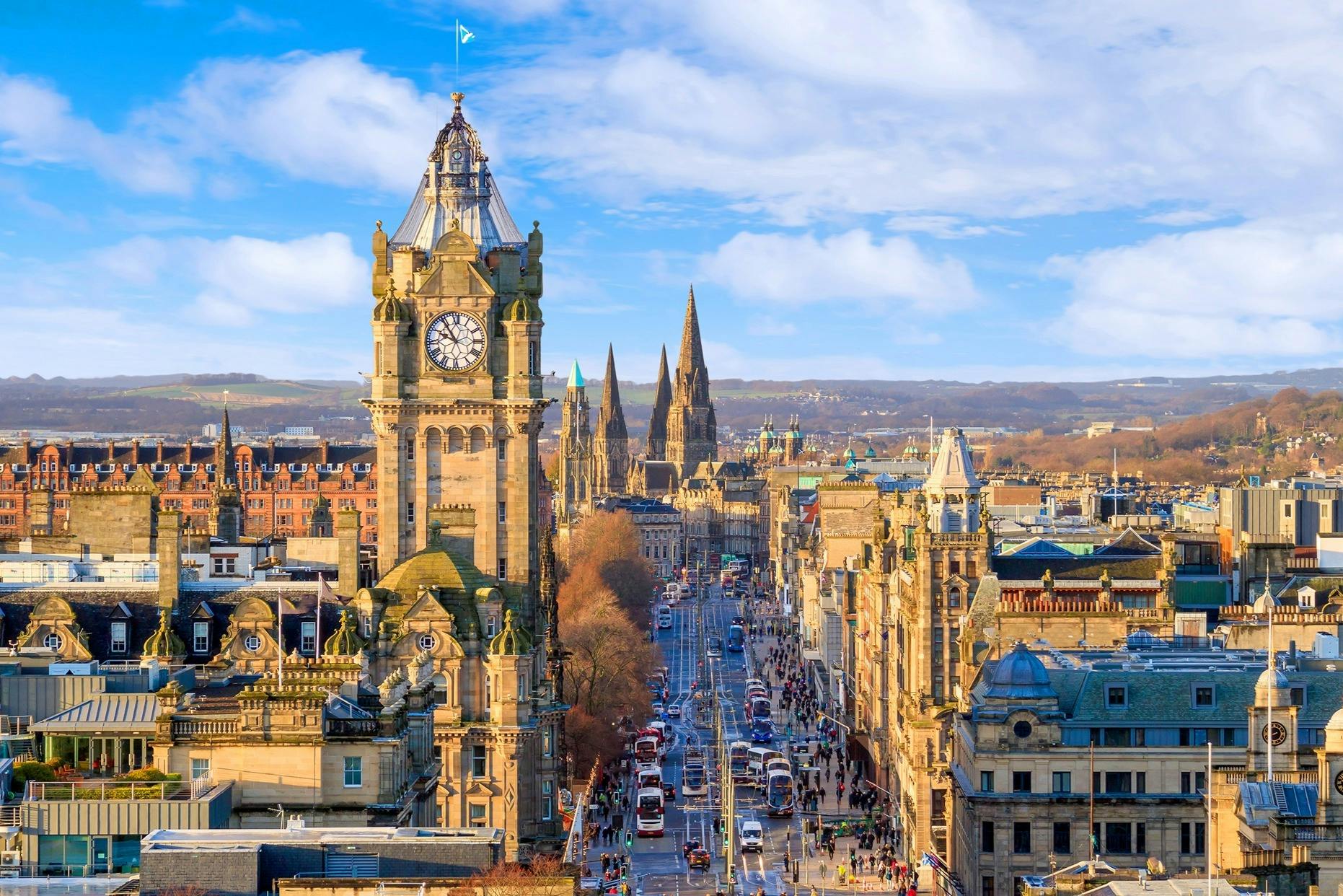 Un paesaggio urbano con una torre dell'orologio, guglie e una strada animata da auto e autobus, sotto un cielo parzialmente nuvoloso.