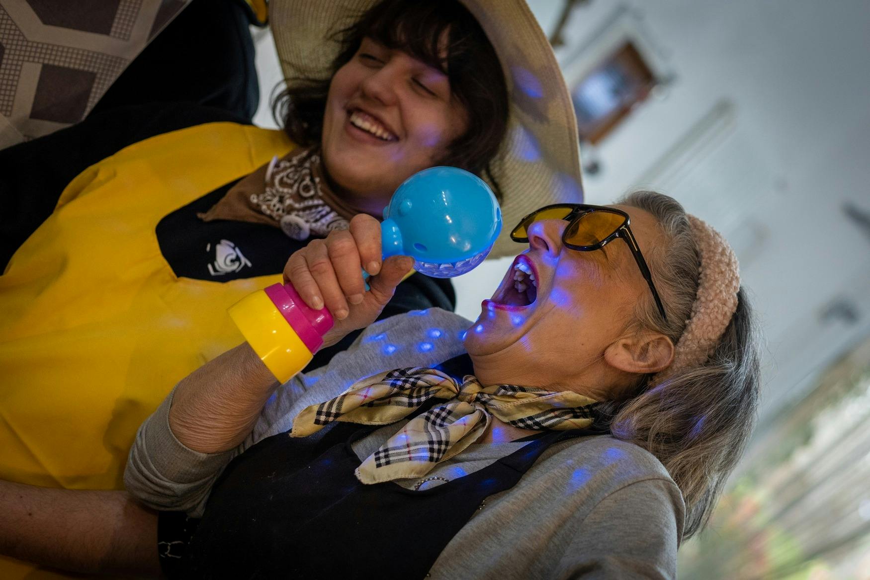 Dues dones gaudint; una canta en un micròfon de joguina, ambdues somrient. Porten barrets i complements de colors.