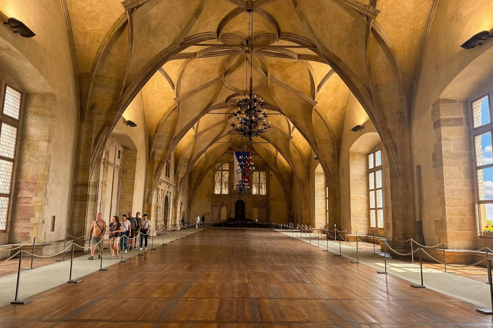 A grand hall with vaulted ceilings, chandeliers, wooden flooring, and people viewing the space along the sides.