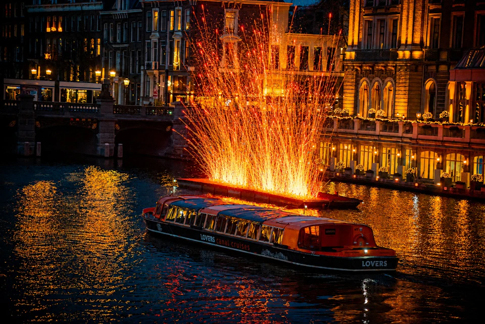 A canal cruise boat passes in front of a fiery light installation on the water at night, surrounded by lit buildings.