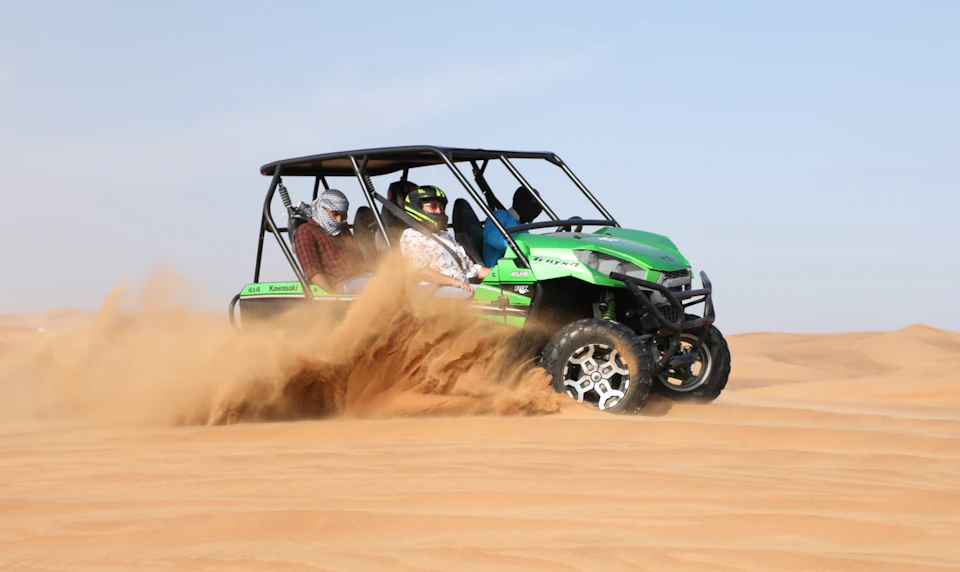 Two people in helmets drive a green off-road vehicle through desert sand, kicking up a cloud of dust.