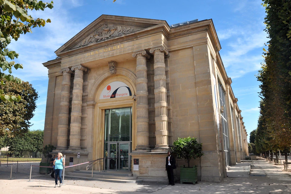 Ornate stone building with four columns at entrance, large arched doorway, sign reading "Musée de l'Orangerie," clear blue sky.