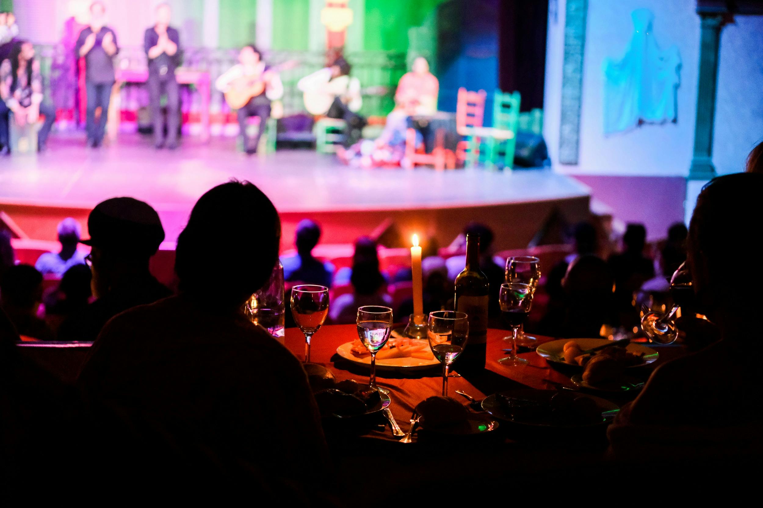 A dimly lit dining area with wine glasses and plates on a table in the foreground, and a stage performance in the background.