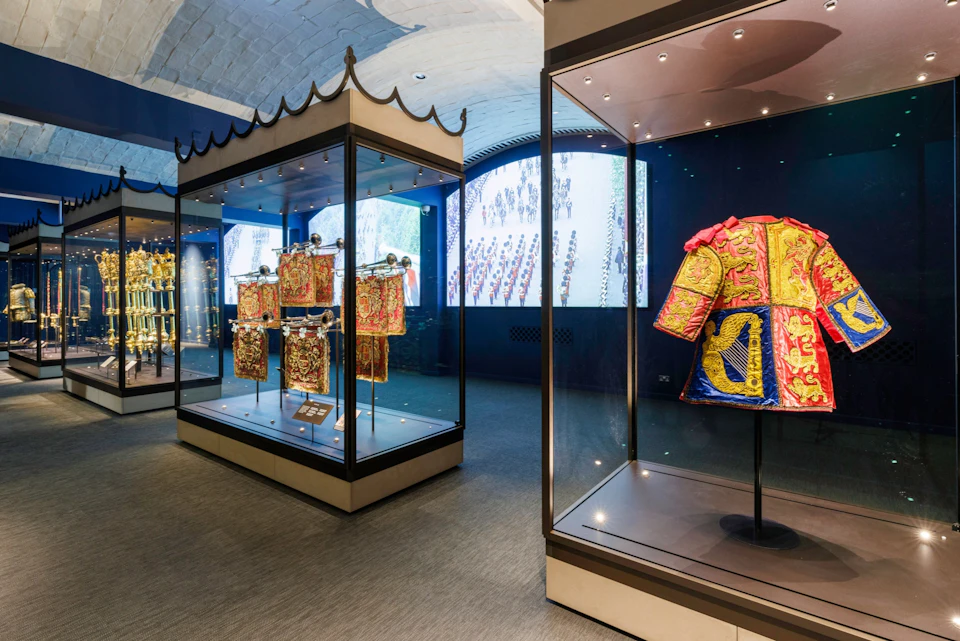 Exhibit with ornate, historical garments in glass cases; screen displaying a parade in the background.