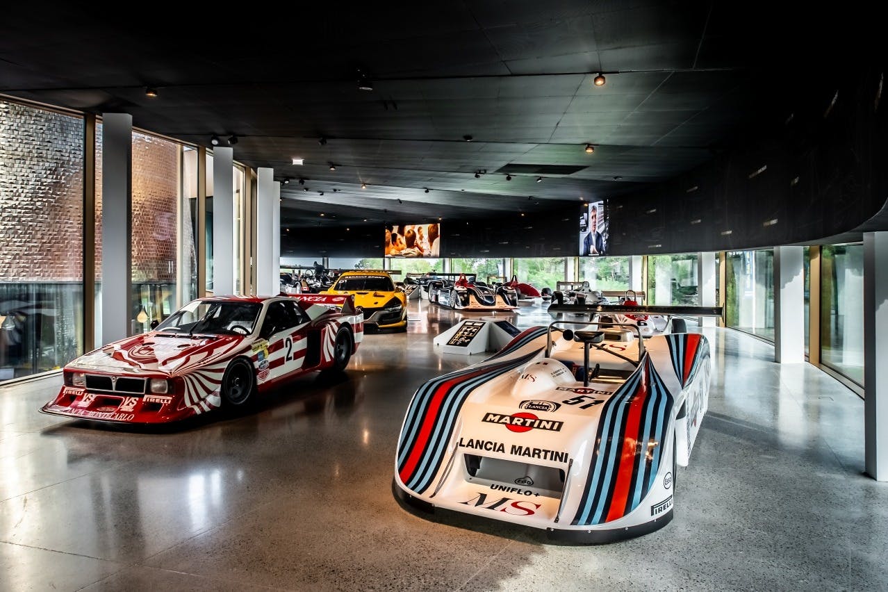 Un elegante showroom automobilistico che espone diverse auto da corsa d'epoca sotto una luce soffusa, tra cui una Lancia Martini in primo piano.