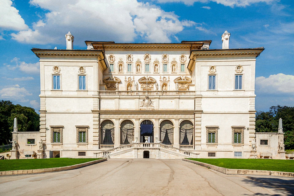 A grand, ornate building with arched windows, statues, and a central staircase, set against a partly cloudy blue sky.
