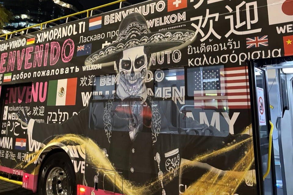 Een bus versierd met een skelet in sombrero en pak, omringd door meertalige welkomstwoorden.