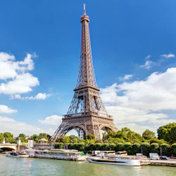 Eiffeltornet med turbåtar dockade längs floden Seine under en blå himmel med spridda moln.
