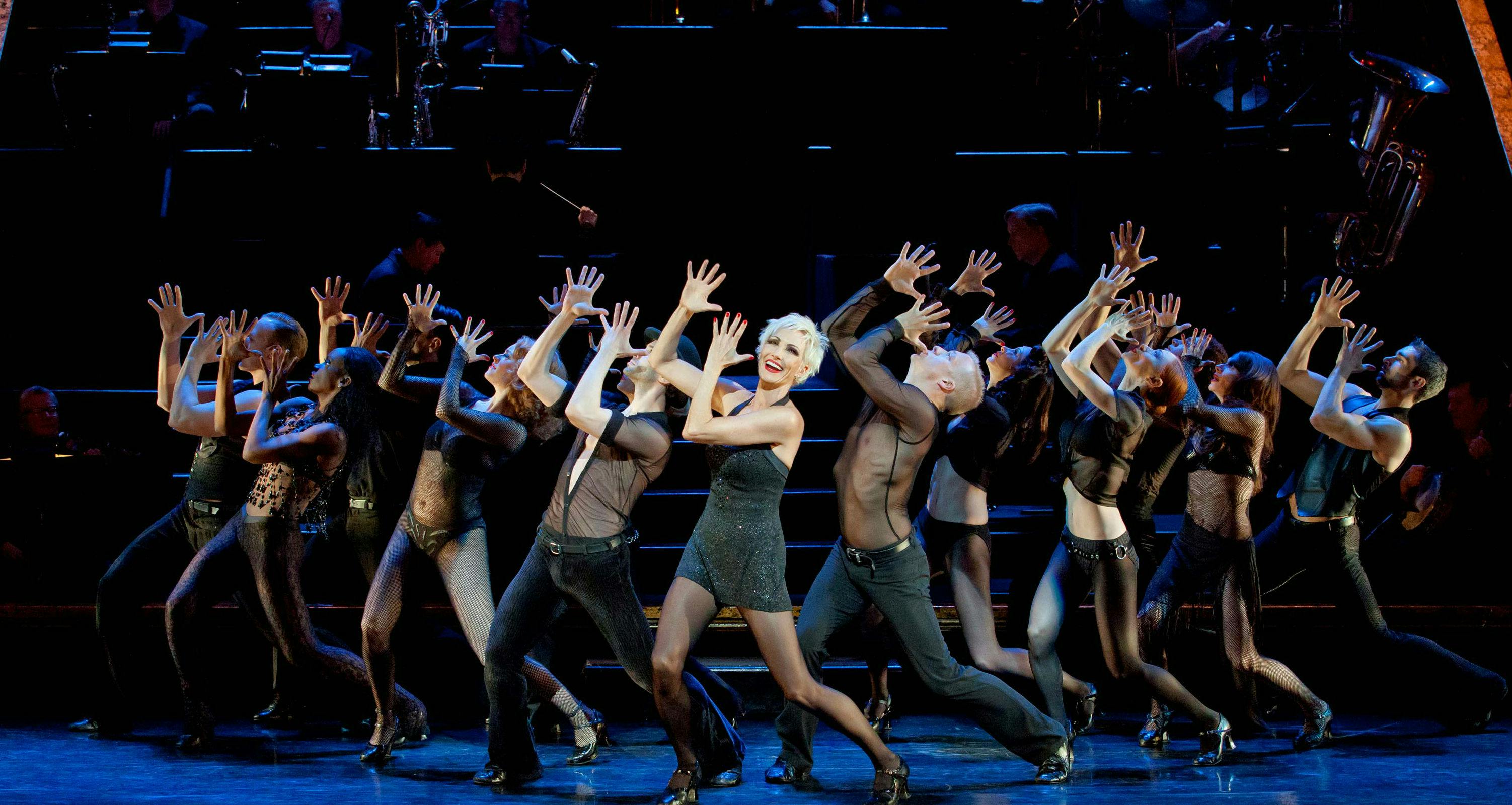 Performers in black outfits dance energetically on stage, hands up, in sync, with a smiling female lead in the center.