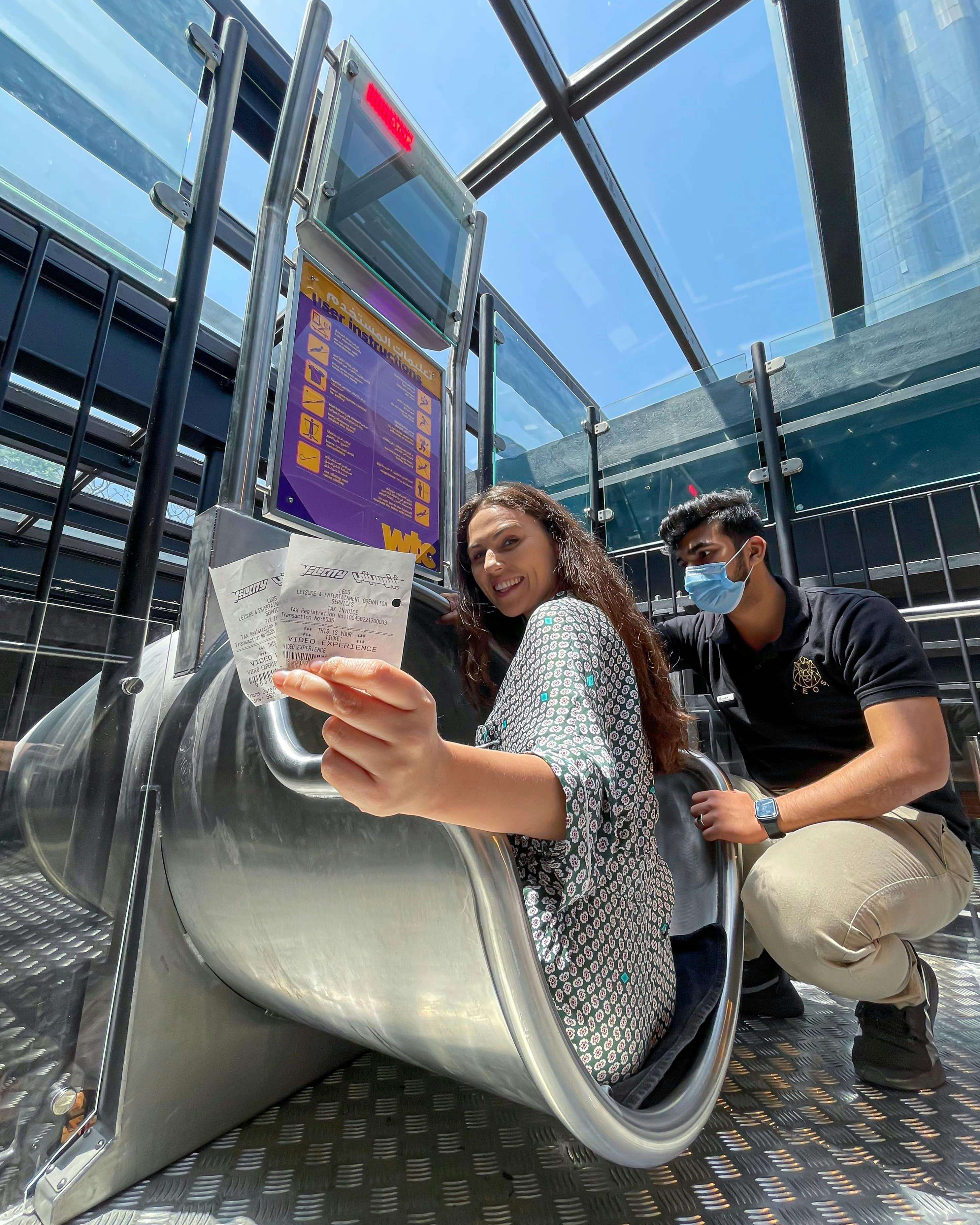 A woman sits at the top of a slide, holding a ticket, while a masked man assists her. They are under a glass roof.