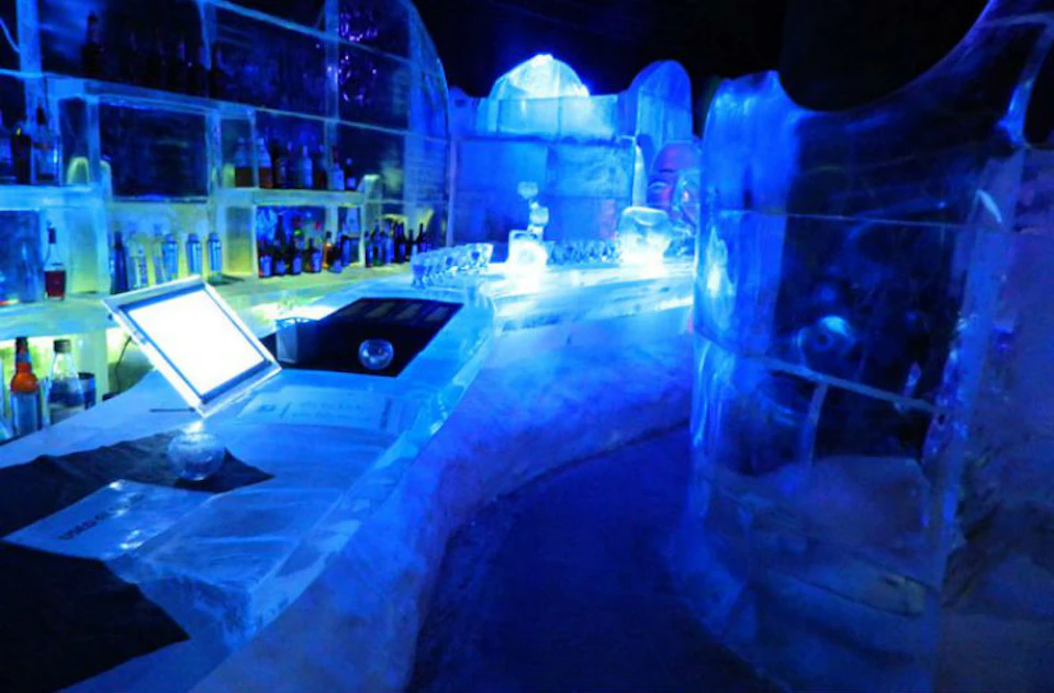 A dimly lit ice bar featuring ice sculptures, a counter, shelves with bottles, and a blue glow from ambient lighting.