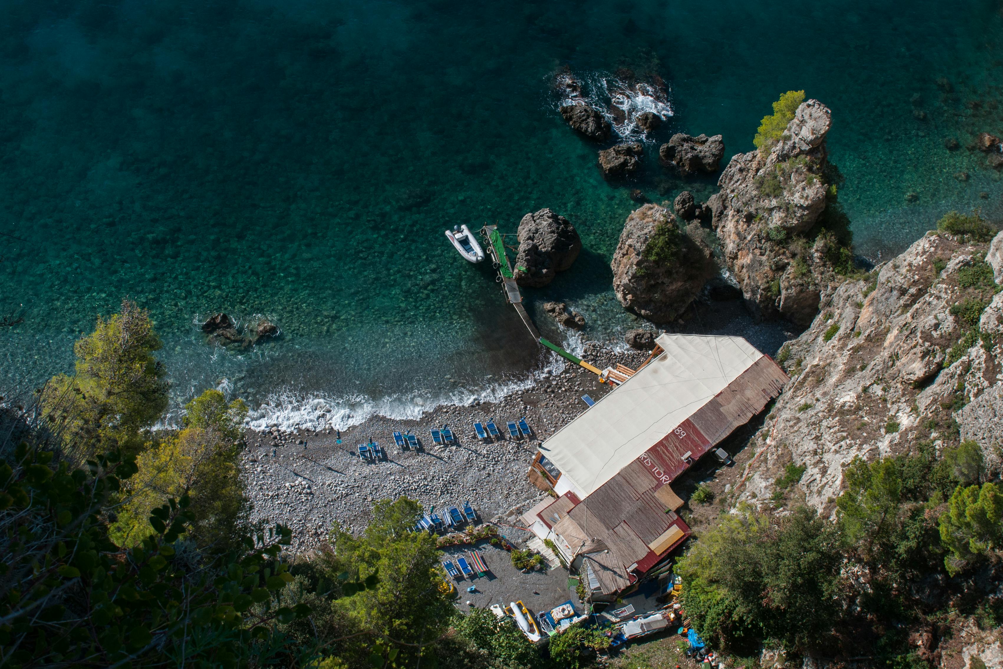 Aerial view of a coastal area with clear turquoise water, rocky shore, beach chairs, a docked boat, and a canopy-covered structure.