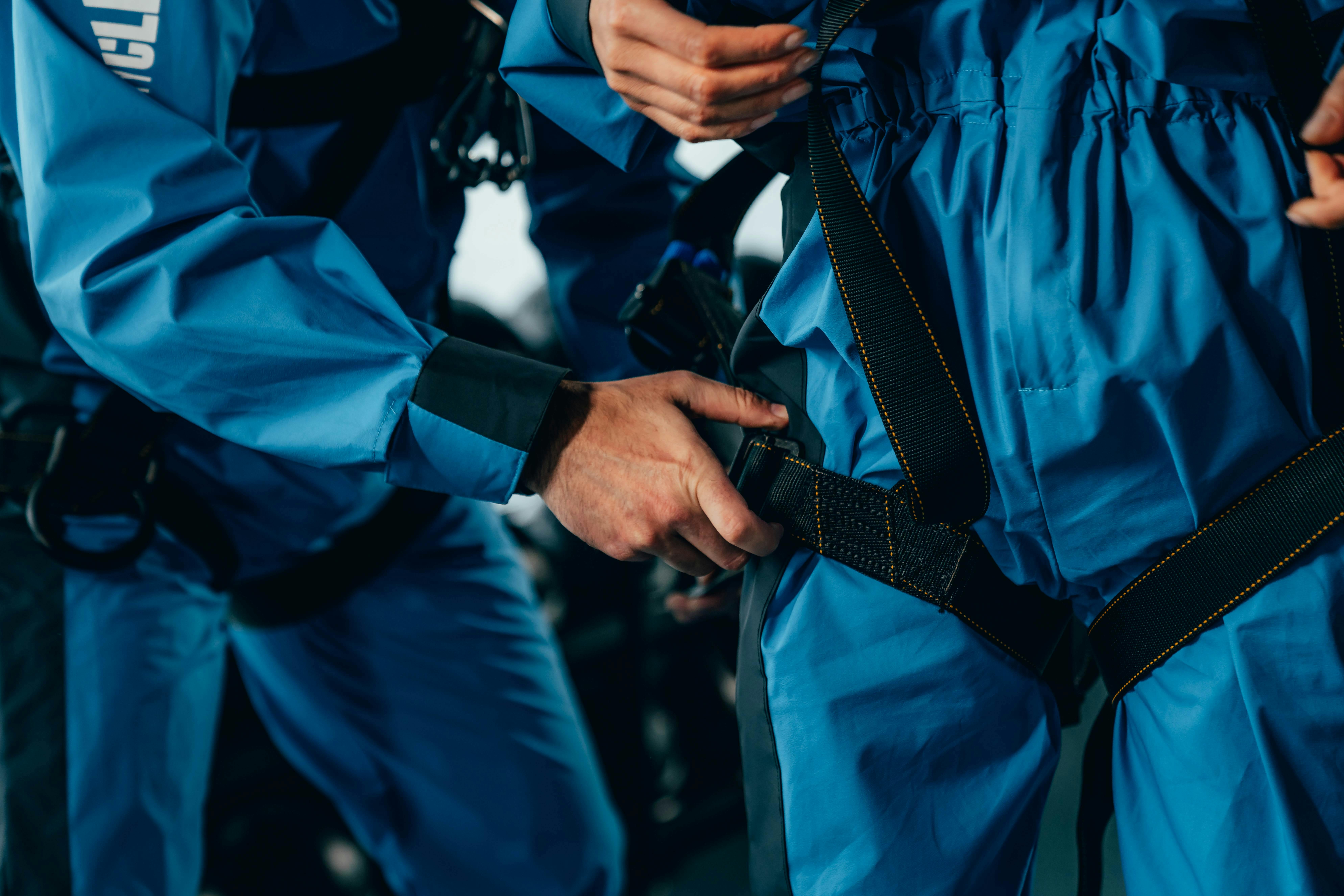 两个穿着蓝色连体裤的人，一个帮助另一个系上黑色安全带扣。