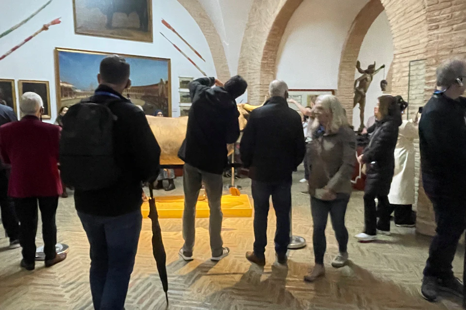 People in a museum viewing a large exhibit, with paintings and sculptures visible in the background.