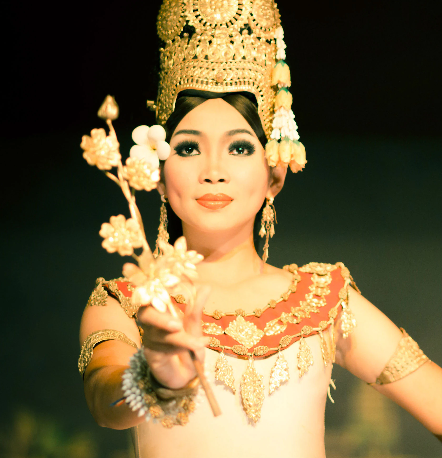 A person wearing traditional gold headgear and decorative attire holds a floral ornament, looking directly ahead.