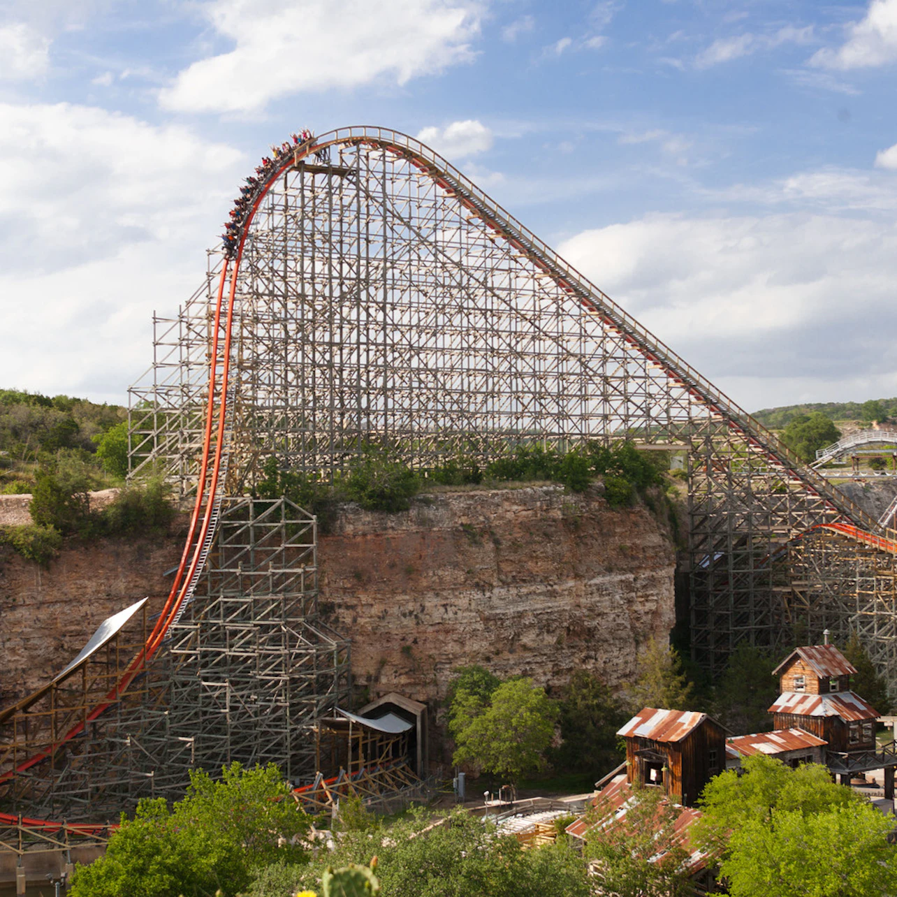 A roller coaster on a steep descent with a wooden support structure, surrounded by trees, cliffs, and rustic buildings.